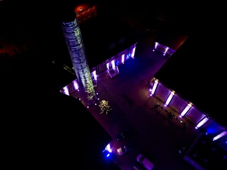 Blick von oben auf einen beleuchteten Platz mit bunten Lichtern und einem hohen Turm.